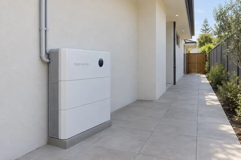 Illustrative render of a stacked home battery on a white rendered wall beside a side path